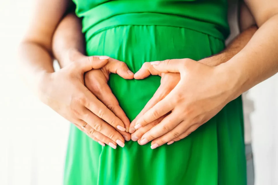 Hände eines Paares formen Herz auf Babybauch einer schwangeren Frau in grünem Kleid.