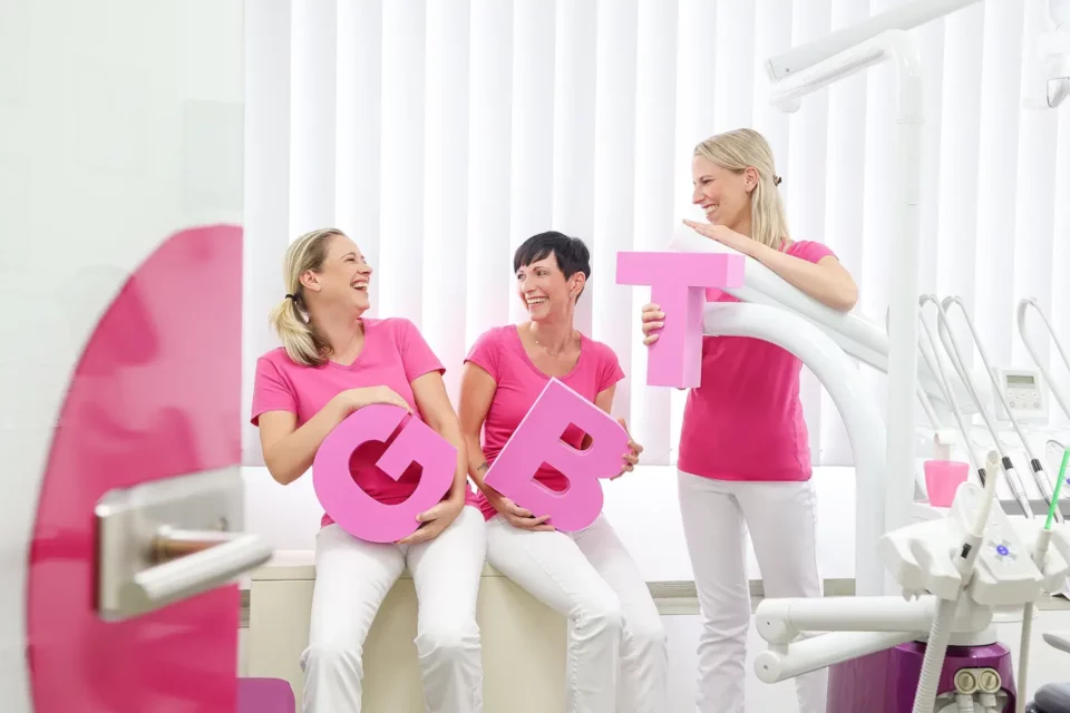 Drei lachende Mitarbeiterinnen in Zahnarztpraxis mit rosa „GBT“-Buchstaben in der Hand.