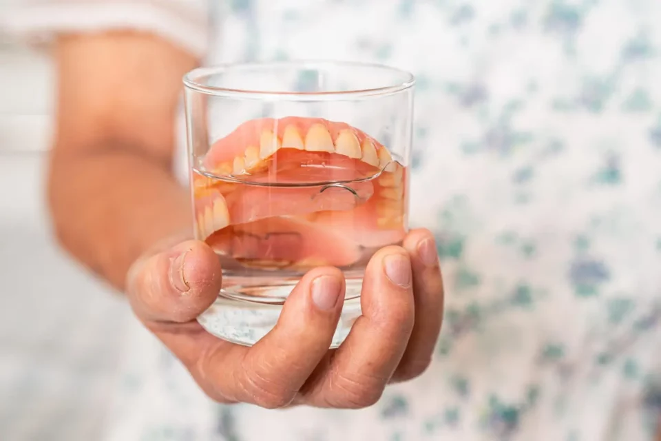 Hand hält Glas mit Zahnprothese in Wasser zur Reinigung und Aufbewahrung.